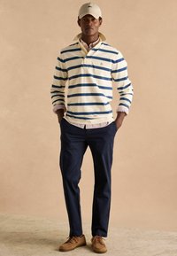 Gestreepte polo met lange mouwen in crème en marineblauw, gedragen over een rood gestreept shirt, gecombineerd met marineblauwe broek en bruine schoenen. Voorzien van een pet.