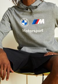 Sudadera gris con los logotipos de BMW y M, texto "Motorsport", capucha con cordón y bolsillo frontal. Usada con pantalones cortos negros mientras está sentado.