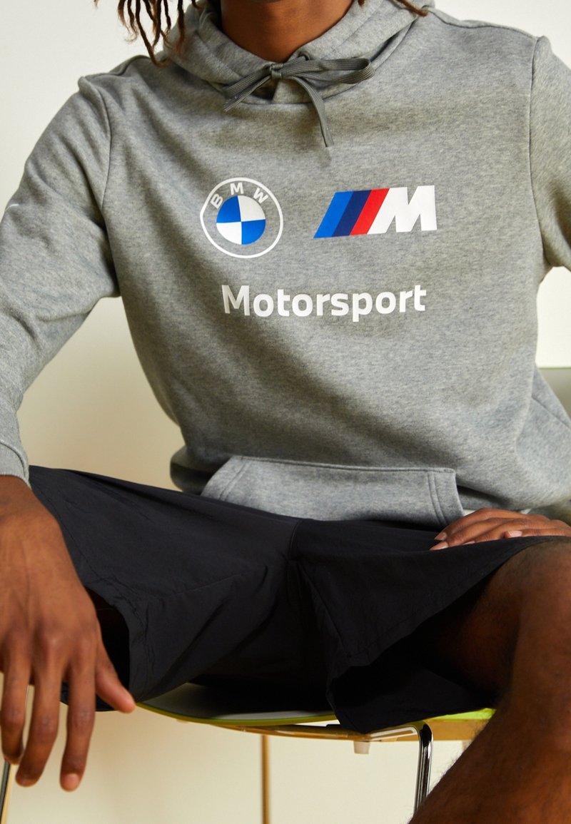 Sudadera gris con los logotipos de BMW y M, texto "Motorsport", capucha con cordón y bolsillo frontal. Usada con pantalones cortos negros mientras está sentado.