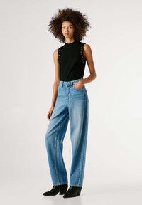 Top negro sin mangas con detalles de ojales en los hombros, combinado con jeans de mezclilla azul claro de talle alto y botas negras de tacón.