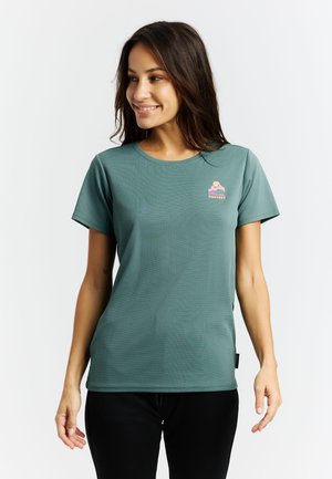 Jonge vrouw die lacht, gekleed in een tealkleurig shirt met korte mouwen en een klein, kleurrijk logo op de linkerborst, gecombineerd met een zwarte broek.