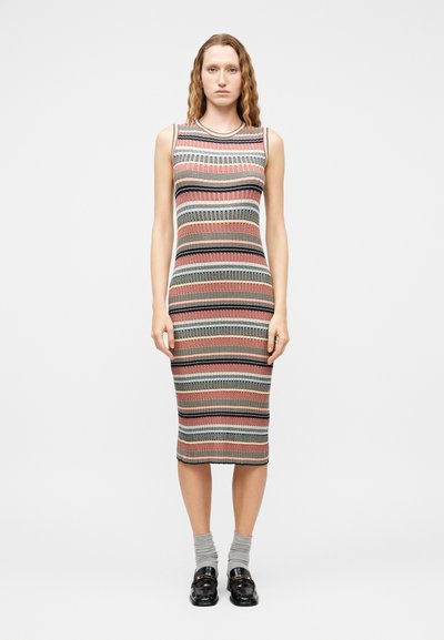 Femme portant une robe en tricot sans manches, mi-mollet, à rayures horizontales multicolores, des chaussettes grises et des mocassins noirs, debout devant un fond uni.
