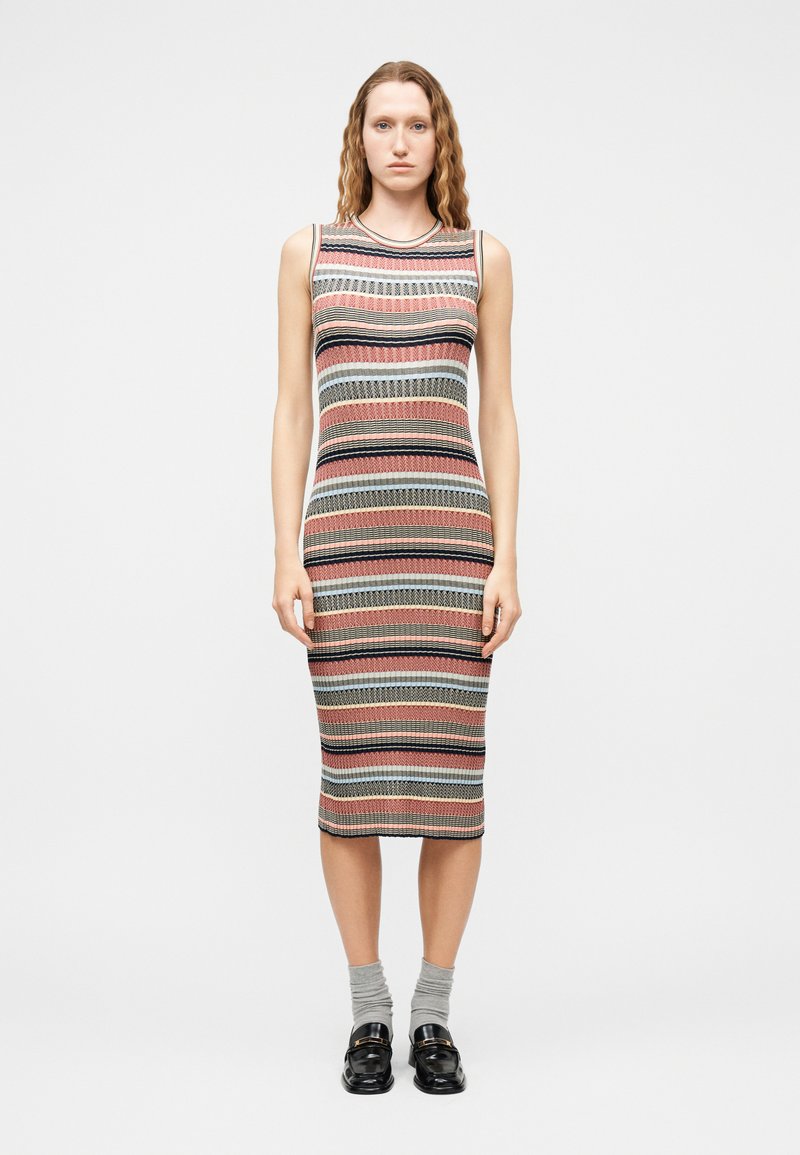 Femme portant une robe en tricot sans manches, mi-mollet, à rayures horizontales multicolores, des chaussettes grises et des mocassins noirs, debout devant un fond uni.