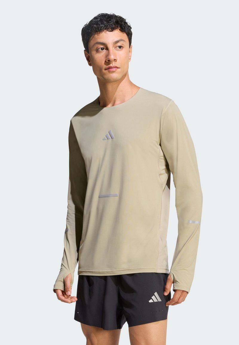 Muž v béžovom športovom tričku s dlhými rukávmi a čiernych šortkách s logami Adidas, stojaci a mierne sa pozerajúci do boku.