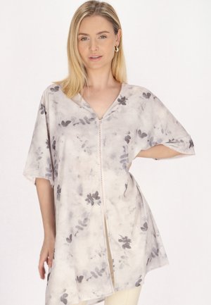 Femeie cu păr blond purtând o tunică lejeră, deschisă la culoare, cu modele florale gri, stând cu o mână pe șold, pe un fundal simplu.