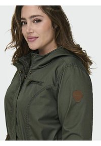 Grüne Kapuzenjacke mit Reißverschluss und Druckknöpfen, mit einer Reißverschlusstasche und einem braunen sechseckigen Logopatch auf der Schulter. Weicher, strukturierter Stoff.