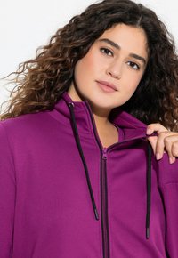 Fialová mikina na zip s černými šňůrkami, s vysokým límcem a hladkou texturou látky. Zip částečně otevřený, odhalující vrstvu pod ním.