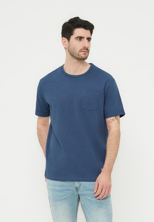 Άνδρας που φοράει μπλε κοντομάνικο μπλουζάκι με τσέπη και ανοιχτόχρωμα τζιν, στέκεται μπροστά σε λευκό φόντο, κοιτώντας προς τα δεξιά.