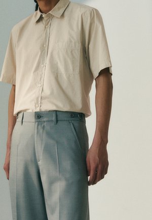 Mann trägt ein kurzärmliges, cremefarbenes Hemd mit Knöpfen, das in hochtaillierte, graue, plissierte Hosen gesteckt ist, vor einem einfarbigen Hintergrund.