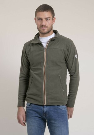 Homme portant une veste polaire zippée vert olive par-dessus une chemise blanche et un jean bleu, debout devant un fond clair uni.