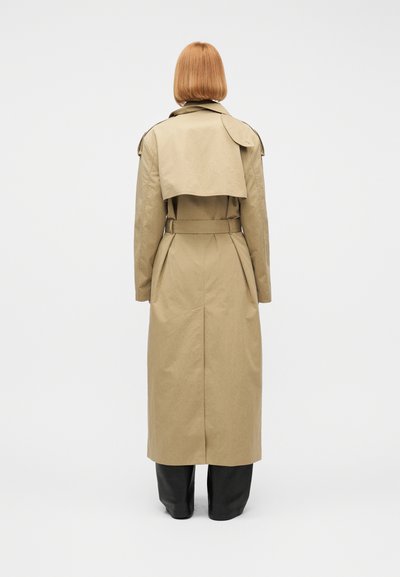 Άτομο με κοντά κόκκινα μαλλιά, φοράει μπεζ καμπαρντίνα και μαύρο παντελόνι, στέκεται κοιτώντας προς ένα απλό λευκό φόντο.