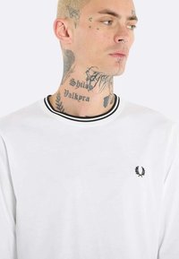 Camicia bianca a maniche lunghe con colletto a costine nero e piccolo emblema nero sul petto. Il tessuto appare liscio e leggero.