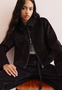 Temno jeans jakna s ovratnikom, vidnim zadrgo in stranskimi žepi. Gladka tekstura z modrimi šivi, v kombinaciji s usklajenimi hlačami.