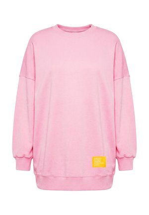 Vaaleanpunainen ylikokoinen collegepaita pitkillä hihoilla ja resorireunuksilla, jossa on pieni keltainen kangasmerkki valkoisella tekstillä helman lähellä.