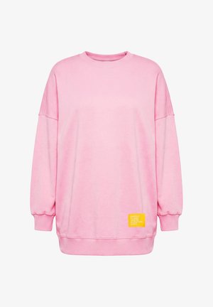 Vaaleanpunainen ylikokoinen collegepaita pitkillä hihoilla ja resorireunuksilla, jossa on pieni keltainen kangasmerkki valkoisella tekstillä helman lähellä.