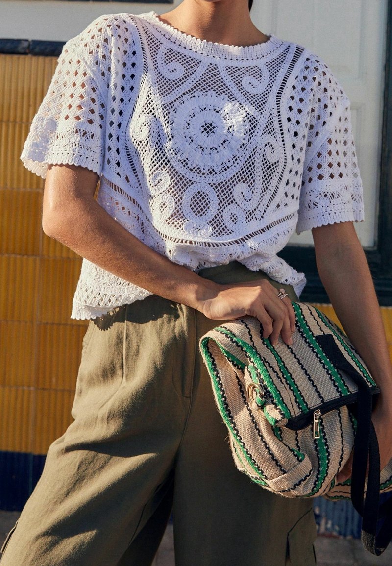 Top de crochet blanco con un escote redondo, que presenta patrones intrincados y mangas cortas. La modelo sostiene un bolso a rayas con detalles en verde.