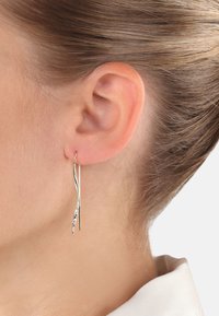 Pendientes de metal dorado con un diseño ondulado, que presentan un acento plateado texturizado. Livianos y alargados, cuelgan justo por debajo de la oreja.
