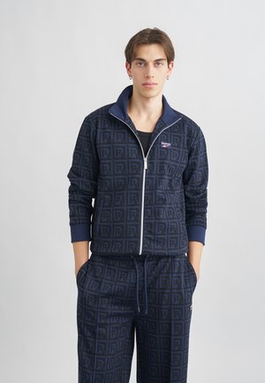 Survêtement marine avec un motif de logo répétitif. La veste a une fermeture éclair à l'avant et un col replié ; le pantalon comprend une taille élastique et des poches.