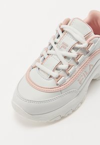 Vita lädersneaker med rosa accenter, tjock sula, rund tå och texturerade detaljer. Har vita snörningar och varumärkeslogotyp på sidan.