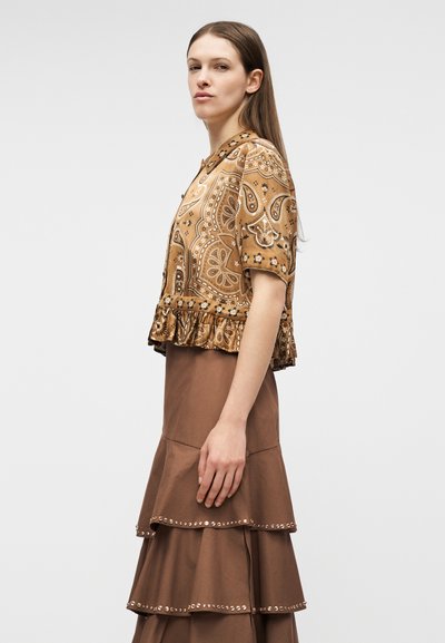 Γυναίκα με καφέ κοντομάνικο μπλουζάκι με παϊένσια και βολάν στη μέση, φορώντας καφέ φούστα με στρωματοποιημένη σχεδίαση και διακοσμητικά καρφιά, όρθια σε προφίλ.