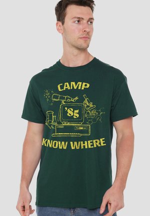 Mann trägt ein dunkelgrünes T-Shirt mit gelbem Design, das einen Vintage-Computer, ein Mikroskop und den Text "CAMP '85 KNOW WHERE" zeigt.