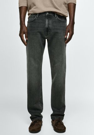 Personne portant un jean noir coupe décontractée, une chemise à manches longues marron clair et des mocassins en daim marron, debout devant un fond uni.