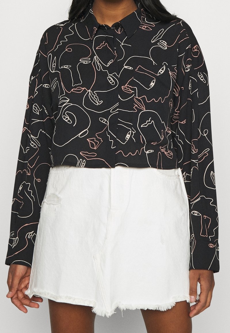 Chemise noire à manches longues et courte avec un motif abstrait de visages en lignes continues, associée à un short en denim blanc à taille haute et bords effilochés.
