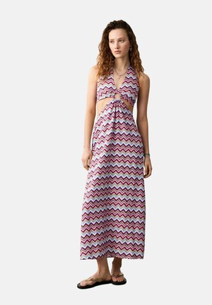 Femme portant une robe maxi sans manches avec un motif zigzag multicolore, des côtés découpés et un détail en anneau au centre devant, assortie à des sandales.