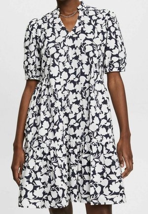 Femme portant une robe à motif floral noir et blanc avec des manches courtes bouffantes, un col en V et une jupe à volants, debout devant un fond uni.