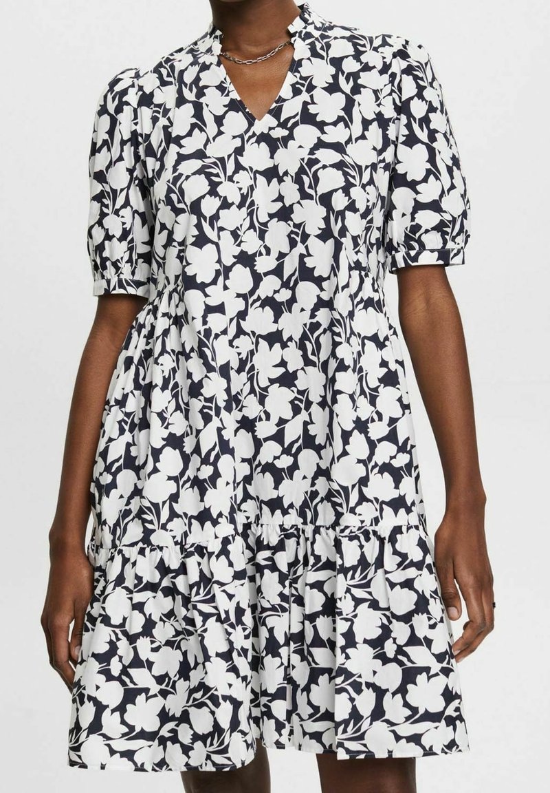 Femme portant une robe à motif floral noir et blanc avec des manches courtes bouffantes, un col en V et une jupe à volants, debout devant un fond uni.