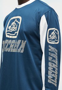 Μπλε μακρυμάνικο πουκάμισο με δίχτυ, λευκά μανίκια, μεγάλο τυπωμένο λογότυπο και υφασμάτινες λεπτομέρειες. Αεριζόμενο ύφασμα με άνετη, φαρδιά εφαρμογή.