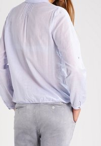 Blouse à carreaux bleu clair avec manches longues, détail froncé au dos et col rond, associée à un pantalon gris clair texturé avec poches.