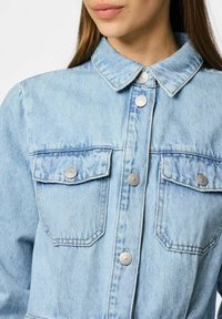 Light blue denim jacket with a collar, silver buttons, and two chest pockets. Features a slightly faded texture and classic design.