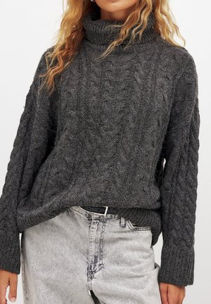 Femme portant un pull à col roulé en maille torsadée gris foncé, rentré dans un jean taille haute gris clair avec une ceinture noire.