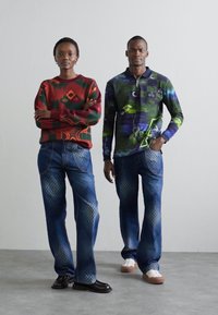 Two models wear vibrant clothing: a red patterned sweater and a green graphic long-sleeve top, paired with blue patterned jeans and shoes.