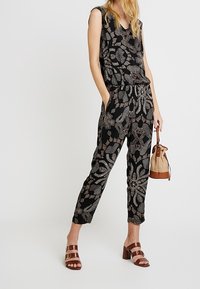 Schwarzer Jumpsuit mit gemusterter Oberfläche, ärmellos und mit schmal zulaufenden Beinen, verziert mit weißen und grauen Blumenmotiven. Kombiniert mit braunen Sandalen mit Riemchen und einer kleinen Tasche.