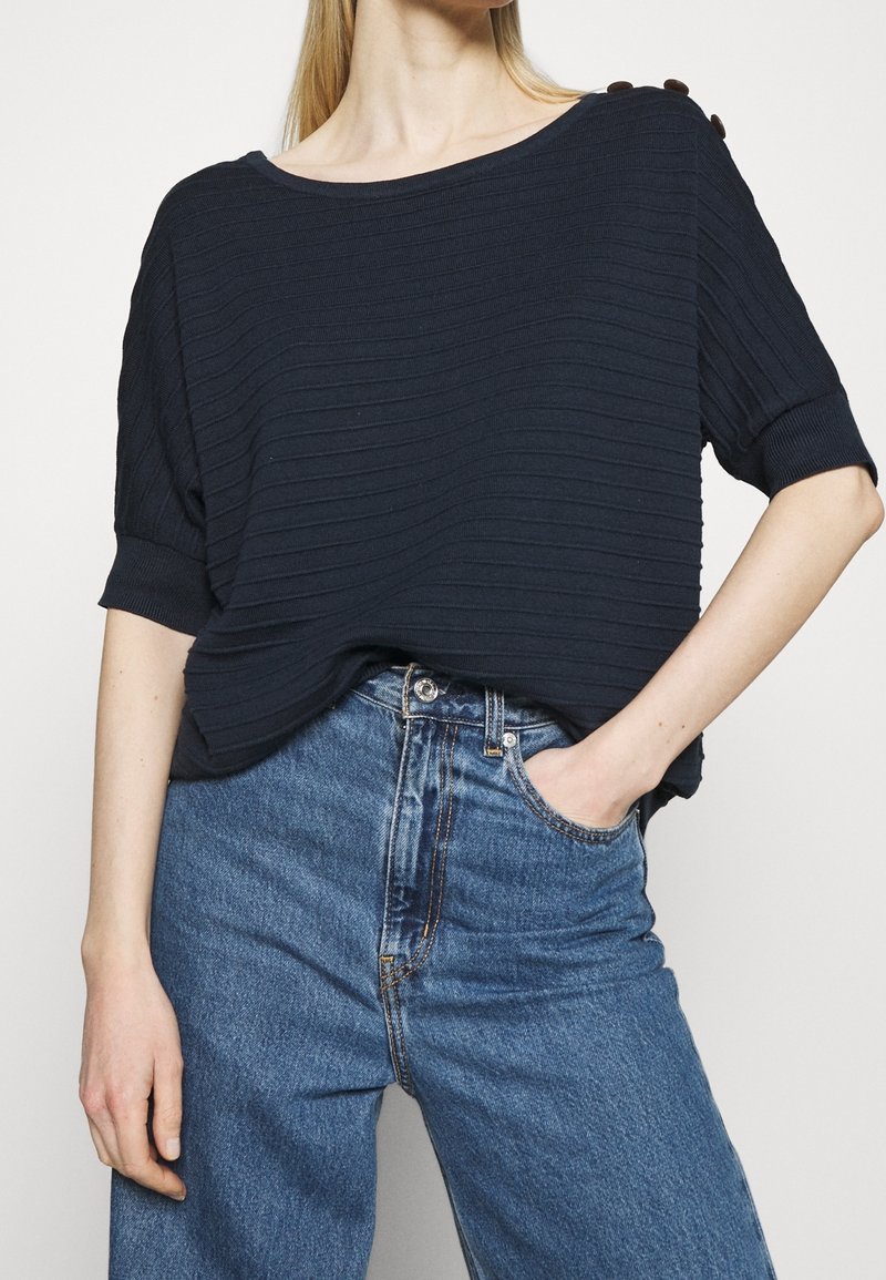 Femme portant un pull à manches courtes côtelé bleu marine avec des boutons sur l'épaule et un jean bleu taille haute, une main dans la poche.