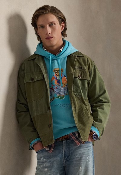 Veste verte portée sur un sweat à capuche bleu avec un motif d'ours, chemise à carreaux en dessous, et jean déchiré avec texture de tissu visible et détail de montre.