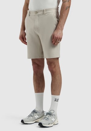 Man wearing beige shorts, white crew socks with black text, and gray New Balance sneakers, standing against a plain light background.