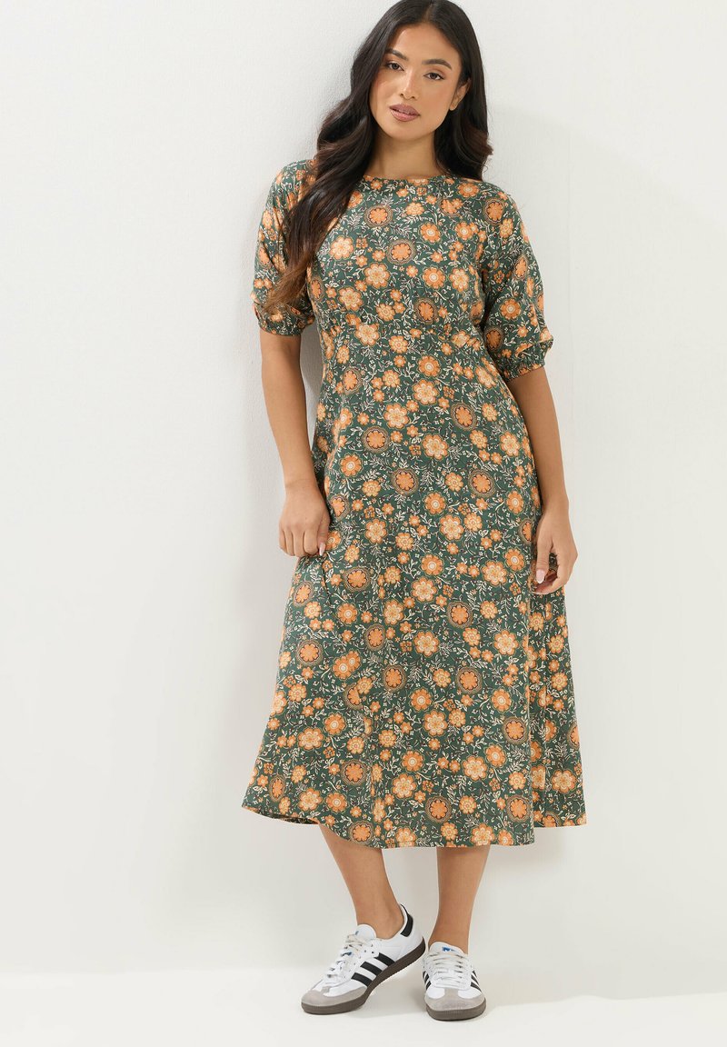 Mujer con cabello largo y oscuro, vestida con un vestido midi verde con estampado floral y zapatillas blancas, de pie frente a un fondo liso y claro.
