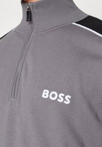 Grauer Reißverschluss-Pullover mit hohem Kragen, schwarz-weißen Schulterstreifen und weißem, gesticktem "BOSS"-Logo auf der Brust.
