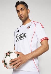 Camisola de futebol branca com detalhes em vermelho, apresentando um logotipo preto no peito. Modelo segurando uma bola de futebol branca com padrão preto.