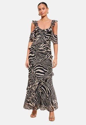 Frau trägt ein Maxi-Kleid mit schwarzem und beigem Zebramuster, Rüschen, schulterfreien Ärmeln und goldenen, überkreuzten High-Heel-Sandalen.