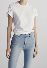 Persoon in een eenvoudig wit T-shirt met korte mouwen en opgerolde mouwen, gecombineerd met lichte hooggaande spijkerbroek, tegen een neutrale achtergrond.