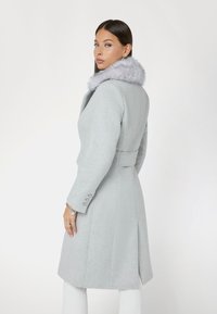 Cappotto di lana grigia con colletto in pelliccia sintetica, design aderente, maniche lunghe e dettagli con bottoni. Texture liscia, lunghezza al ginocchio, con una silhouette sartoriale.