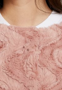 Růžová umělá kožešina s měkkým, strukturovaným povrchem zdobeným vířivým vzorem; na pozadí je bílý límec košile.