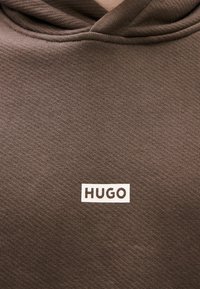 Rudas džemperis su gobtuvu, turintis gofruotą tekstūrą ir baltą stačiakampį logotipą su užrašu „HUGO“ viduryje priekyje.