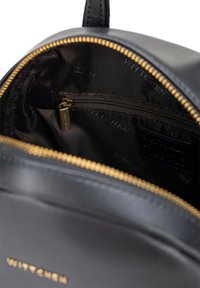 Vnitřek černého koženého batohu se zlatým zipem, obsahující malou kapsu na zip a tmavou látkovou podšívku s logem.