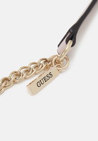 Catena in metallo dorato con un'etichetta rettangolare incisa con la scritta "GUESS" in nero. Il manico è in pelle nera e rosa con una finitura lucida.