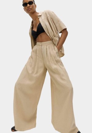 Kvinde iført beige bukser med vide ben og matchende kortærmet skjorte over en sort crop top, med sorte solbriller og hænder i lommerne.